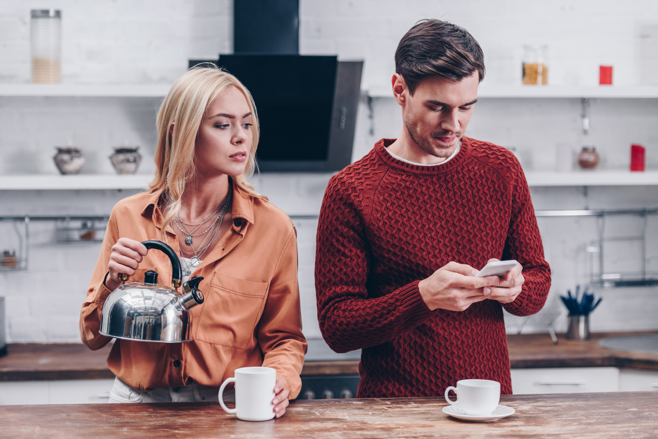 jealous young woman with kettle looking at husband with smartphone in kitchen