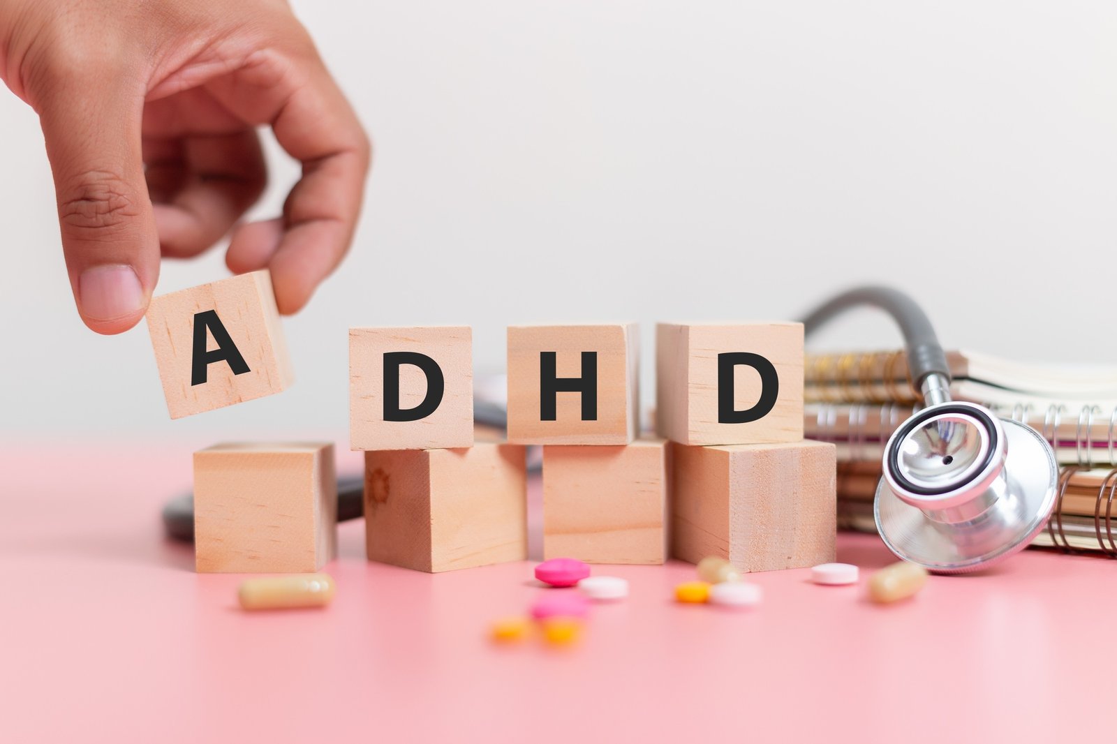 Abbreviation of ADHD on wood cube block on note book with Stethoscope and pills on pink table.
